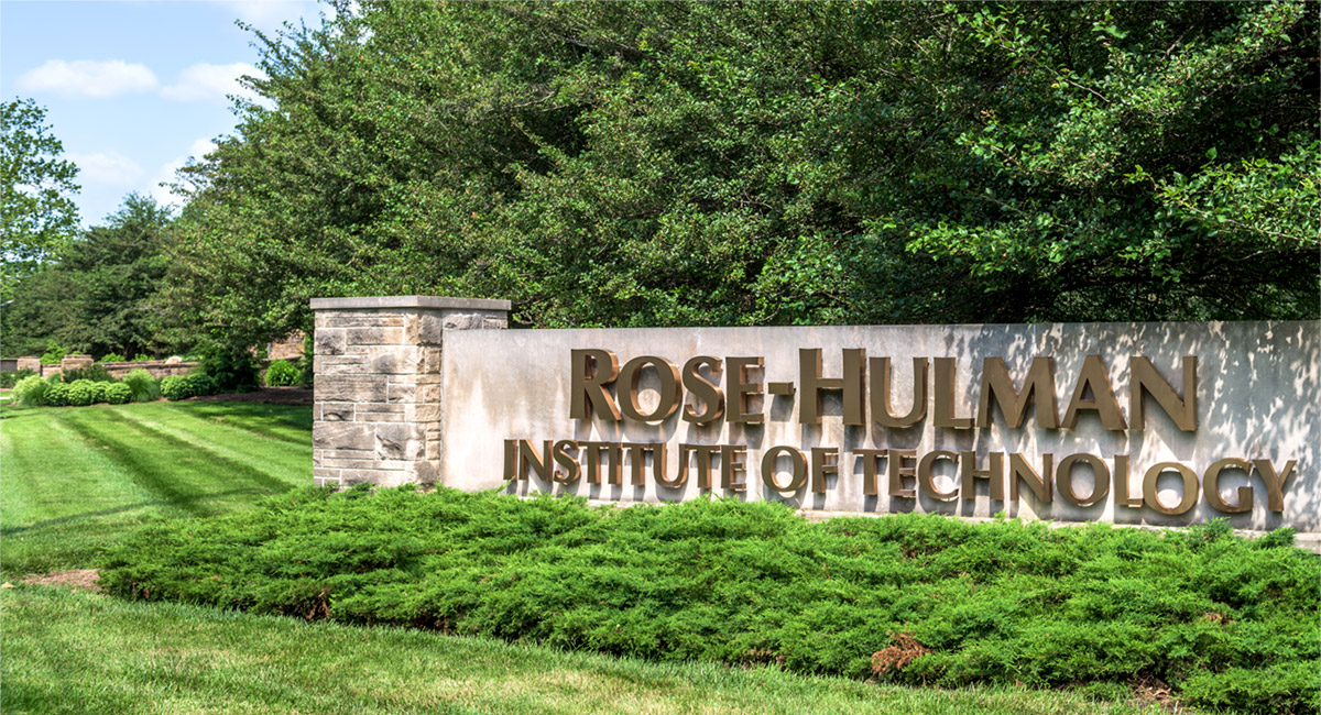 Image shows the stone and concrete sign with gold letters reading Rose-Hulman Institute of Technology at the entrance to the campus. It is couched in green leaves above and green bushes and grass below and to the side.