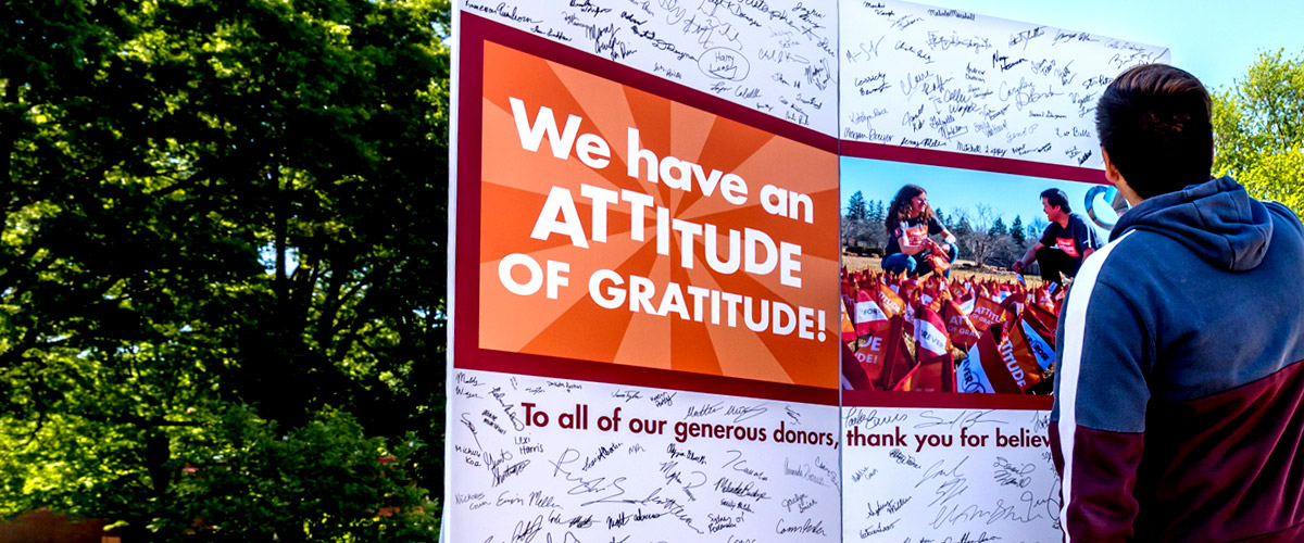 Image shows a male student reading a double poster-board Attitude of Gratitude card, which has been signed by students showing their gratitude for donor support of the institute. The card is on display outside with a large leafy tree in the background.