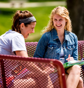 Image shows two female Rose-Hulman students laughing while sitting on a bench on campus on a sunny day.