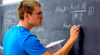 !Male student working a math problem on a chalkboard.