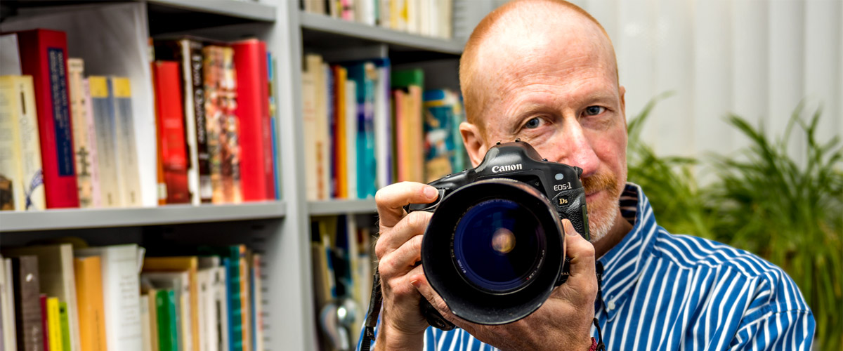 Spanish Professor John Gardner stands with a large camera inside his office.