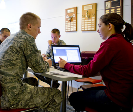 Man in military uniform sits beside a female and her laptop.