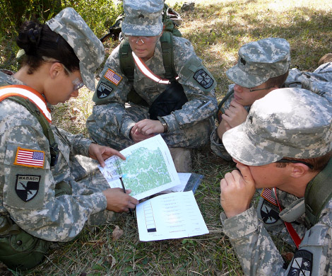 Group of 4 individuals in uniform sit in a circle on the grass looking at documents.