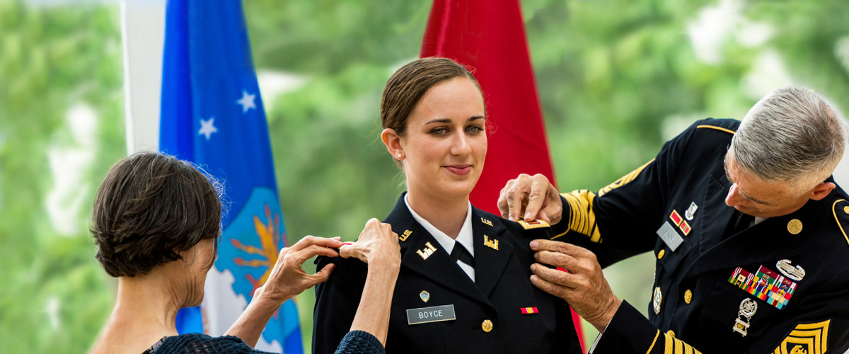 Female in uniform is joined by a male on one side of her and female on the other side of her, who add pin to her uniform.