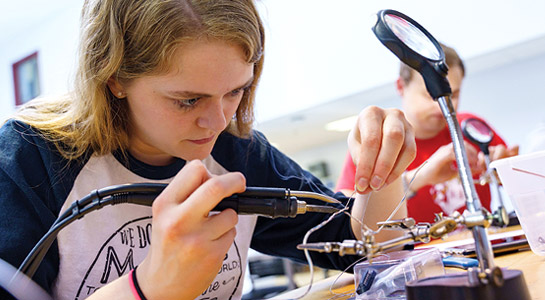 !Female student uses electronic equipment to assemble a student project.