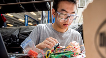 Male student wearing goggles works on wiring for electrical device. 