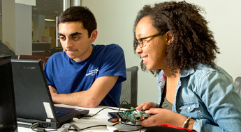Female student holds circuit board and looks at laptop computer screen with male student. 