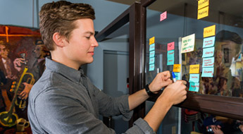 Male student uses Post-It notes to organize ideas on a pane of glass.