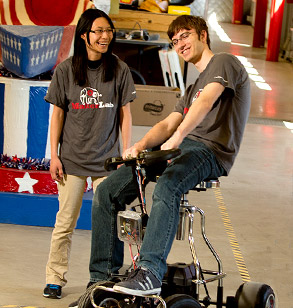 Male student rides single-person electric vehicle in the Branam Innovation Center as laughing female student looks on.
