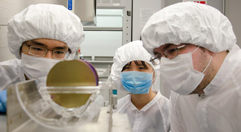 Three students in clean room-style hair covers, face masks, and lab coats examine items in the laboratory.