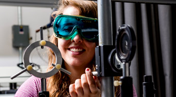 A female optical communications student wearing safety goggles and conducting an experiment using sophisticated optical communications equipment.