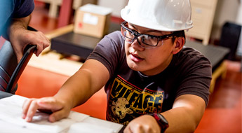 Male engineering student wearing safety glasses and a hardhat reviewing construction plans.