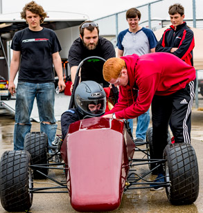 One student drives an open-wheeled Formula SAE car while others make adjustments and look on.