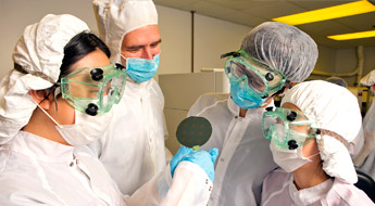 Person in clean room attire uses and instrument to handle a metal disc.
