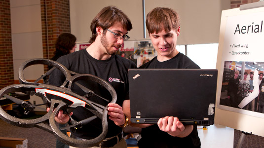 A male student holds an aerial drone while another student holds a laptop computer during a poster presentation.