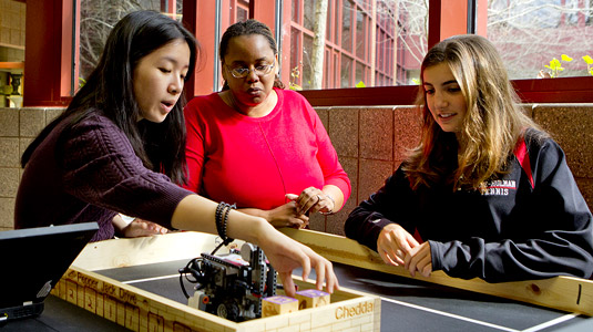 Two female students and a female instructor converse while standing at a table with a small robot and robotics course on top.