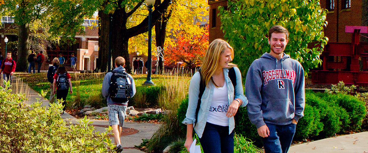 Multiple students walk outside on campus. The leaves on the trees are multiple colors –green, yellow, and orange.