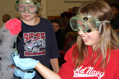 Smiling young female student wearing lab goggles and gloves touches flower.