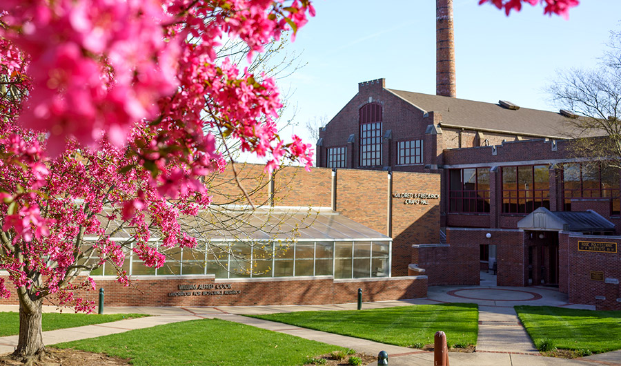 Exterior of Crapo Hall showing green grass and the greenhouse in the foreground and under a blue sky.