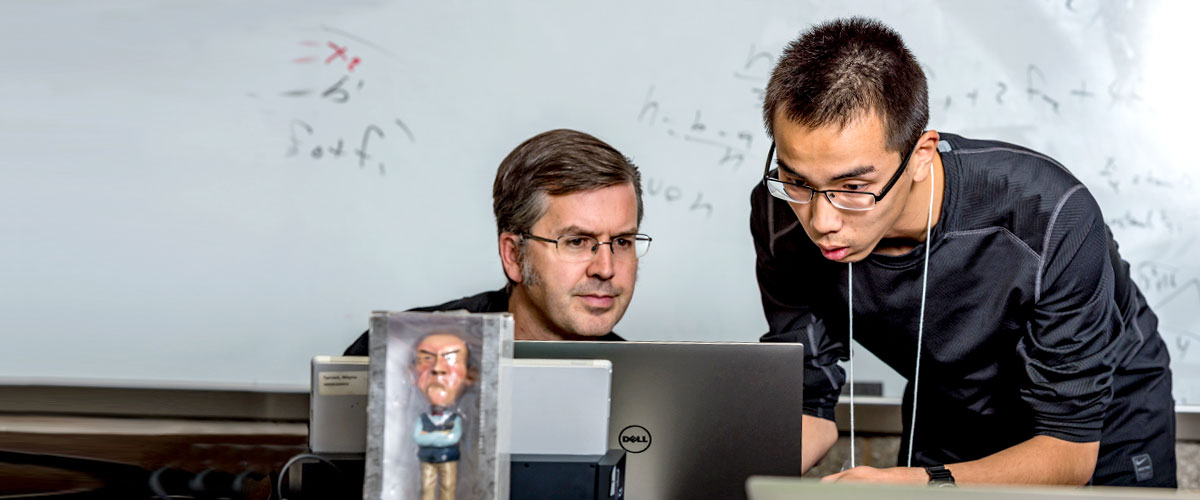Student works on a problem at his professor’s desk as the professor looks on.
