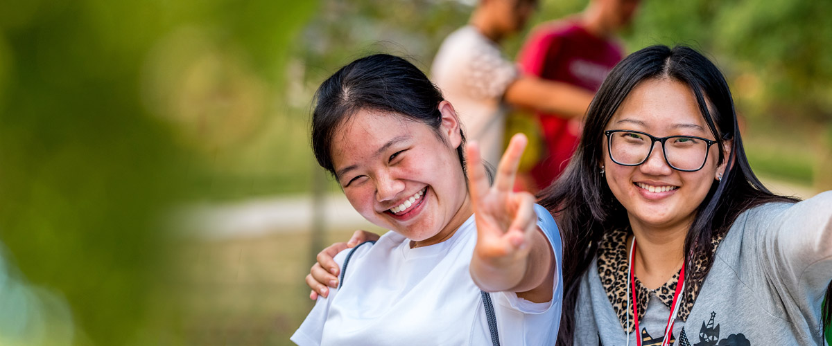 Two female Asian international students smiling and posing together for a photo. One is making the peace sign. 