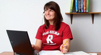 !Image shows a female student smiling and looking off to her right while wearing a red Rose-Hulman T-shirt and taking notes as she uses her laptop computer.