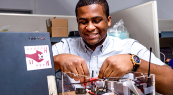 Image shows a male student working on an electronics project and smiling in a lab on campus.