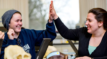 !Image shows two female students high-fiving each other and smiling as they work on a project together in a biology laboratory.