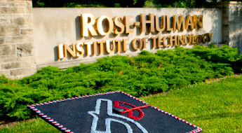 !Image shows the large stone and concrete entrance sign for Rose-Hulman. The sign has large gold-colored letters reading Rose-Hulman Institute of Technology. The sign is lighted by bright sunlight and sits on green grass with low green bushes directly in front of the sign