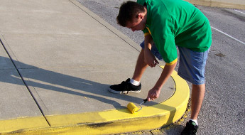 Image shows a male student using a paint roller to paint a yellow “no parking” curb on a street somewhere on campus.