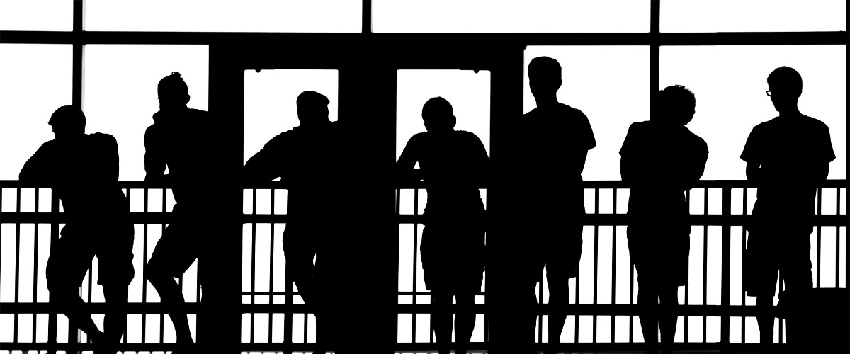 Image shows silhouettes of students leaning on a guard rail in front of doors and large windows