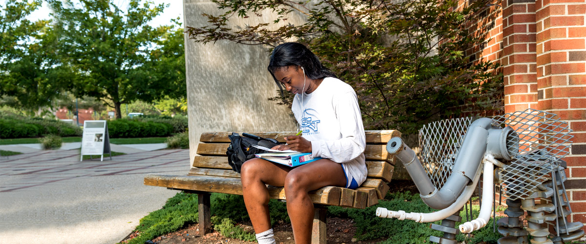 emale student works on homework seated on a bench outside of Moench Hall on a sunny day.