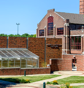 !Exterior of Crapo Hall showing green grass and the greenhouse in the foreground and under a blue sky.