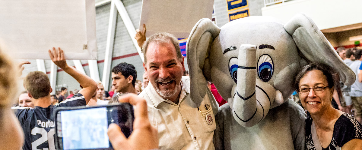 Image shows a student shooting a photo of her smiling parents standing arm-in-arm with Rosie, the Rose-Hulman elephant mascot. They are inside a busy Sports and Recreation Center.