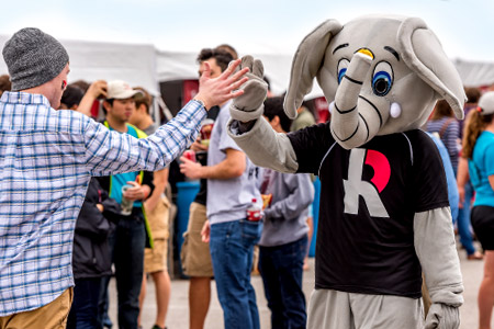 Rosie, the Rose-Hulman mascot, stands and gives high fives to passing students. Rosie is a smiling elephant. 