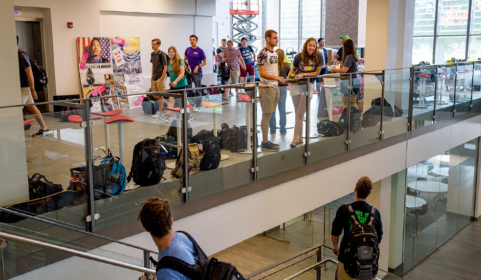 One student standing on a stairway, talking to two students leaning over glass partition above in the student union.