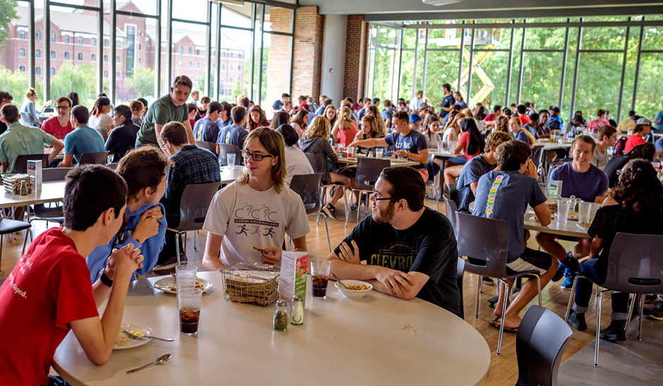 A group of students seated around a round table in the dining room with large windows in the background.