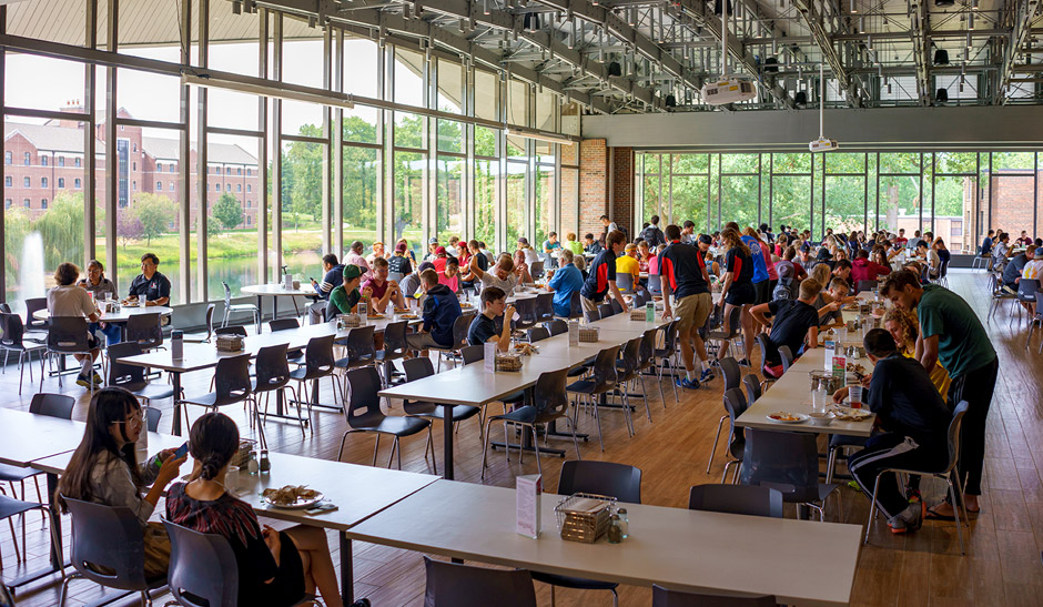 Dining area full of students talking and eating with large windows and lake view in background