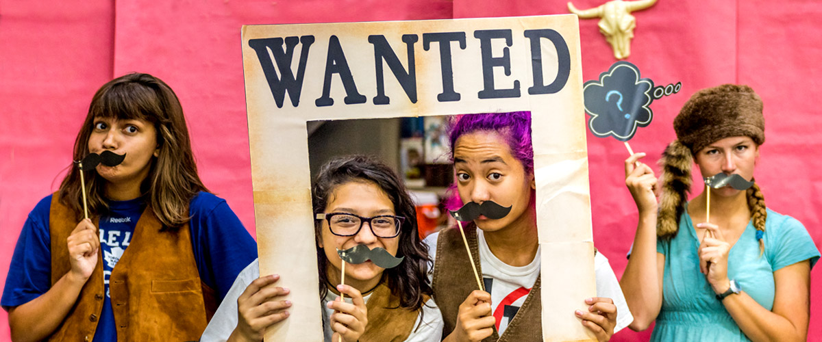 Four female students posing in an old West setting, each holding a fake mustache under their noses and looking very serious (except for one student, who is smiling). Two are standing inside the frame of a 'Wanted' sign.