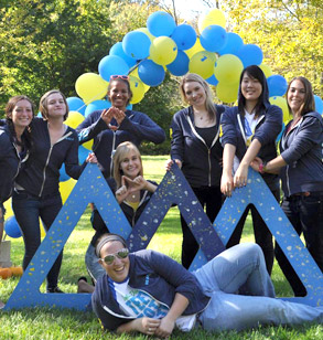 Eight smiling members of the Tri-Delta sorority pose with three large blue delta symbols on a green lawn.