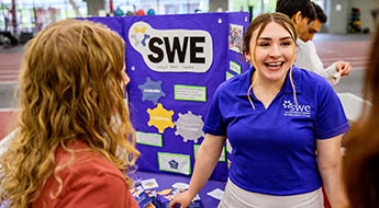 Female students stand beside Society of Women Engineers display board.