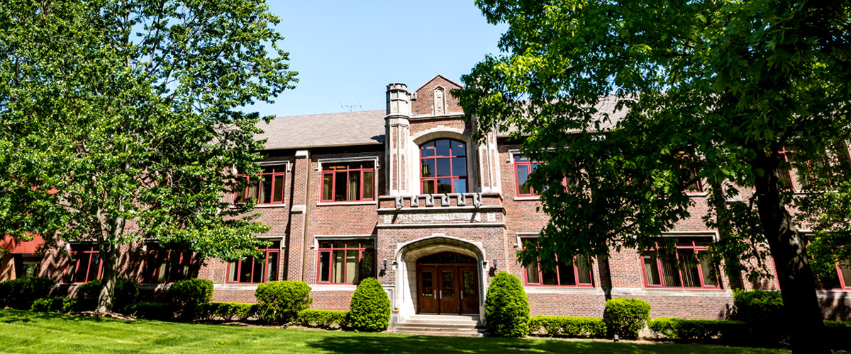Image shows Rose-Hulman’s stately Hadley Hall framed by blue sky, leafy green trees, and green grass.