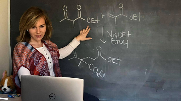 Rebecca DeVasher standing in front of a chalk board displaying chemical formulas and looking at her computer to teach her online class.