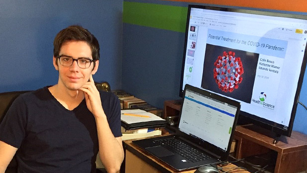 Colin Beach smiling at his desk with a model of the coronavirus shown on his computer screen.