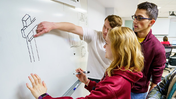 Three mechanical engineering students working on a project together and drawing on a white board.