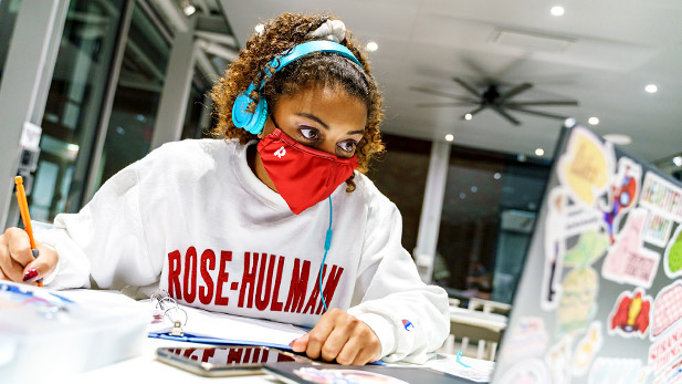 Student wearing a mask working at her laptop computer in a campus common area.
