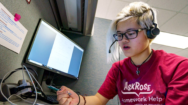Image shows student employee of AskRose talking on the phone while consulting a textbook to help a student caller.