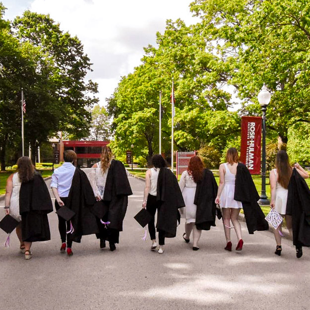 Image shows from-behind-view of several graduating seniors walking up the main road onto campus holding with their commencement regalia with Hadley Hall in the distance.