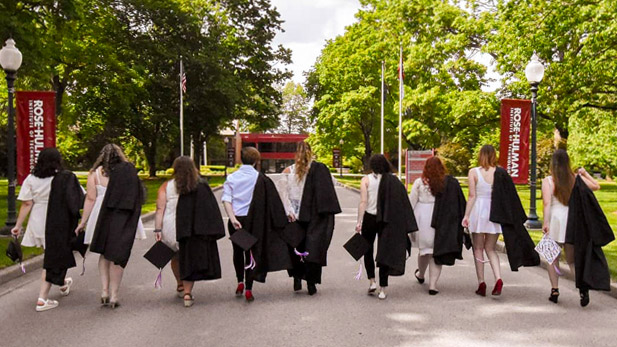 Image shows from-behind-view of several graduating seniors walking up the main road onto campus holding with their commencement regalia with Hadley Hall in the distance. 