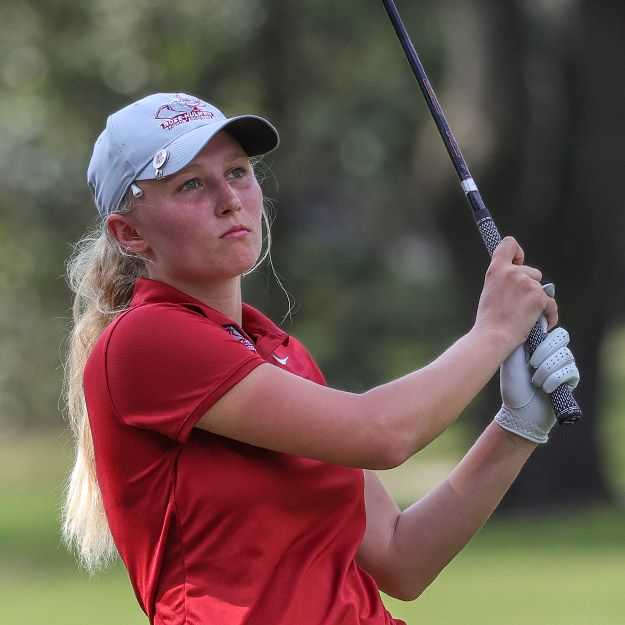 Erin Nolan, wearing a red Rose-Hulman golf shirt, a black golf skirt, and a Rose-Hulman hat over her blonde ponytail, swings her club on the course.